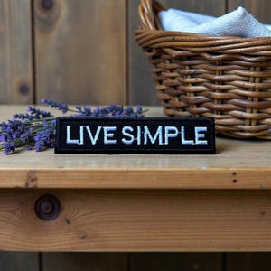 以下が含まれることがあります： 「LIVE SIMPLE」の白い文字が刺繍された黒い長方形のパッチ。木製の表面に表示され、背景にはラベンダーと柳のバスケットがあります。