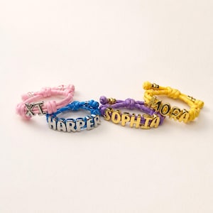 May include: Four bracelets made of colorful cord with metallic letter beads. One pink bracelet features the letters XI, a blue bracelet spells HARPER, a purple bracelet spells SOPHIA, and a yellow bracelet spells MORA in gold-toned letters.