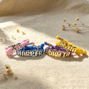 Puede incluir: Cuatro pulseras de cordón de colores con cuentas de letras metálicas. Una pulsera rosa deletrea XI, una azul HARPER, una morada SOPHIA y una amarilla NORA. Cada pulsera presenta un diseño anudado con cuentas metálicas integradas en el cordón.