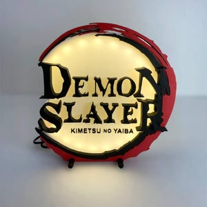 Puede incluir: Un letrero redondo e iluminado con las palabras "Demon Slayer" en letras negras estilizadas. El letrero tiene un borde rojo y negro y el texto "Kimetsu no Yaiba" debajo. Está iluminado con pequeñas luces de tono cálido.