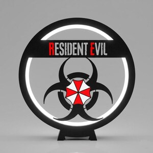 Peut inclure: Logo Resident Evil noir et blanc avec un symbole de danger biologique et le texte "RESIDENT EVIL" en rouge et blanc. Le logo est circulaire avec une bordure lumineuse blanche et une base noire.