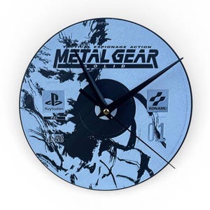 May include: A round wall clock featuring the Metal Gear Solid video game design. The clock face is a light blue with black graphics and text, including the PlayStation and Konami logos. Black clock hands indicate the time.