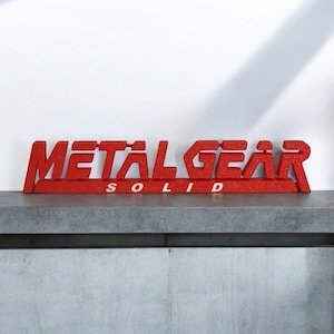 May include: Red glittery "METAL GEAR SOLID" sign. The sign is displayed on a gray surface against a white background. The letters are bold and stylized, creating a striking visual effect. The sign is a decorative item.