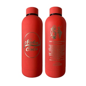 Puede incluir: Dos botellas de agua rojas aisladas con tapas negras. Una botella presenta el logotipo "Nuka Cola" en dorado, mientras que la otra tiene texto y un gráfico en dorado. El texto dice "THE TASTE OF THE USA SINCE 1844".