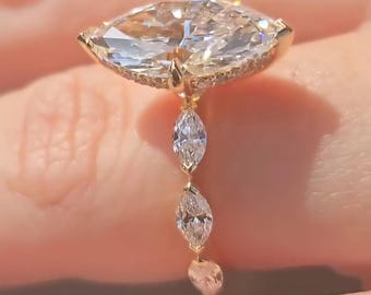 May include: A gold ring featuring a large, marquise-cut diamond as the centerpiece, surrounded by smaller stones. Three dangling, pear-shaped diamonds hang below the main stone, adding a delicate touch to the design. The ring is set against a soft, blurred background.