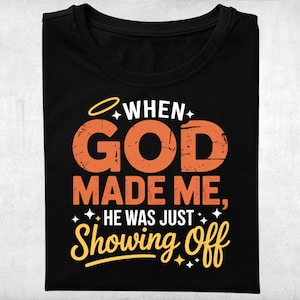 Op de afbeelding: Zwart T-shirt met de tekst "WHEN GOD MADE ME, HE WAS JUST SHOWING OFF" in oranje en geel. Het ontwerp bevat een halo en sterren. Het T-shirt is netjes opgevouwen.