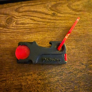 Op de afbeelding: Zwart en rood 3D-geprint gereedschap met het woord "Padawan" erop. Het gereedschap heeft een ronde rode knop en een rood, rechthoekig stuk dat ervan uitsteekt. Het gereedschap ligt op een houten oppervlak.