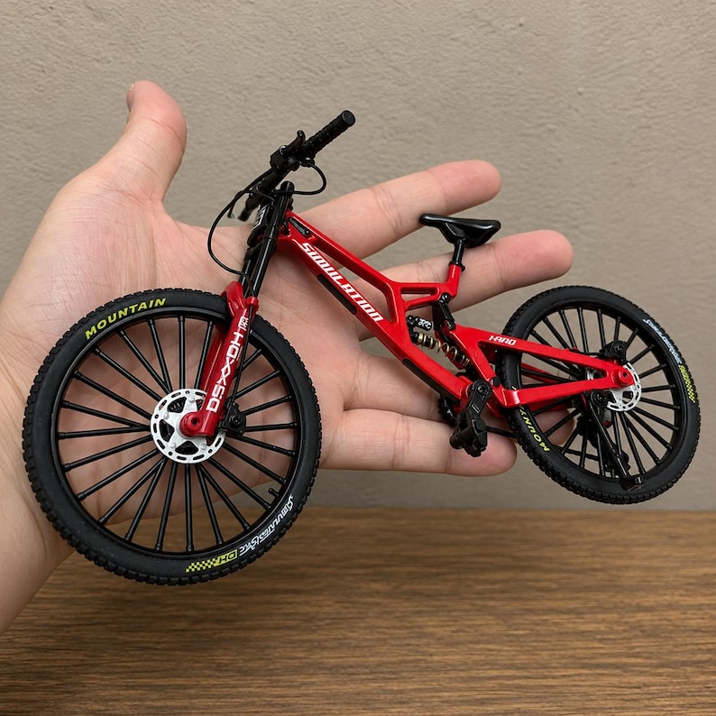 May include: A miniature red mountain bike model featuring black spoked wheels with yellow lettering, a rear suspension coil, disc brakes, and the word SIMULATION printed in white on the frame. The bike includes black handlebars and a black seat.