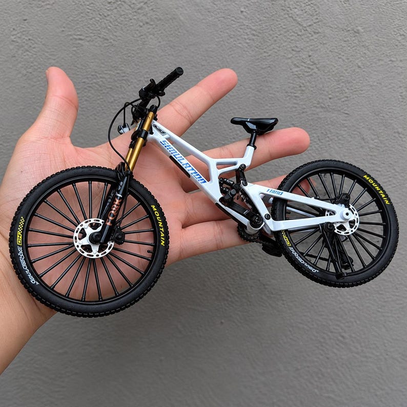 May include: A miniature white mountain bike model with black tires, gold-toned front suspension forks, and black handlebars. The frame features blue text reading SIMULATION and MOUNTAIN on the tires. It includes disc brakes and a black seat.