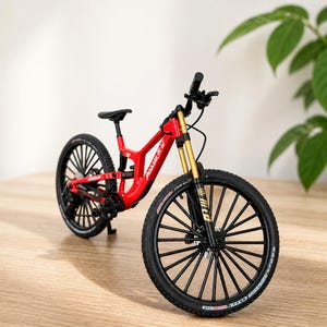 Pode incluir: Modelo de bicicleta de montanha vermelha e preta com garfos de suspensão dourados. A bicicleta tem pneus pretos e um selim preto. O modelo está numa superfície de madeira.