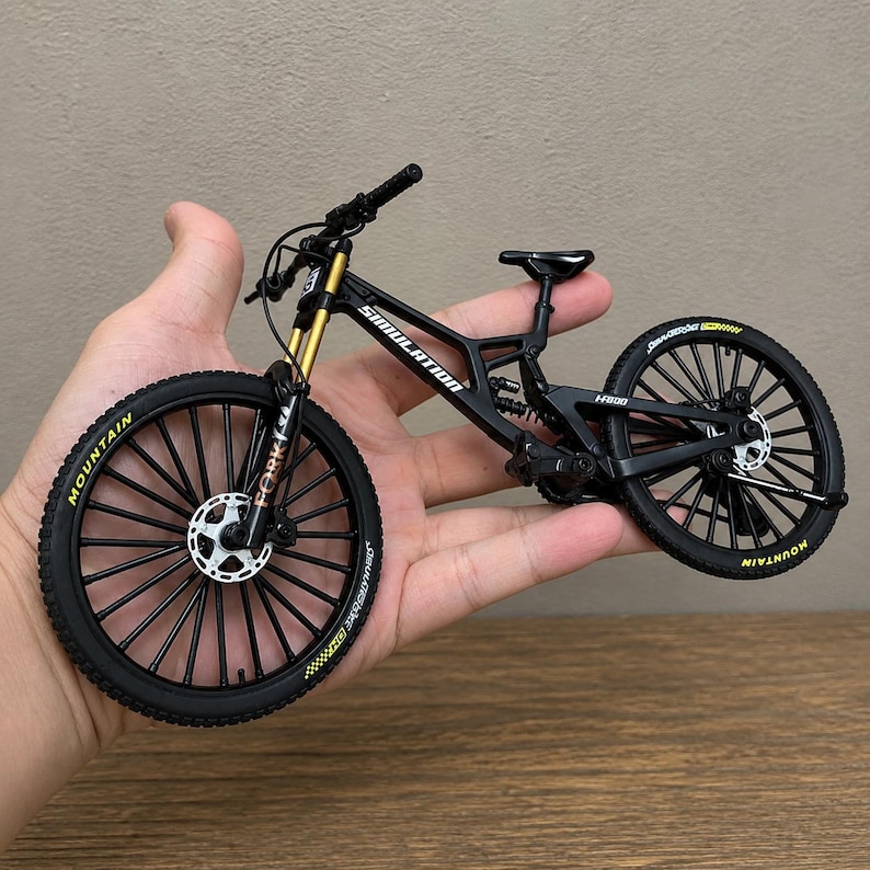 May include: A miniature black mountain bike model featuring gold-colored front suspension forks, black spoked wheels with yellow lettering on the tires, a rear shock absorber, and the word SIMULATION printed in white on the frame's top tube.
