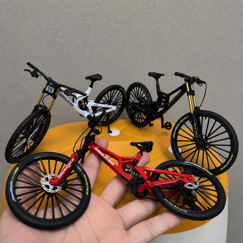 May include: Three miniature mountain bike models in red, white, and black. Each bicycle features a detailed frame, thin black spoked wheels with yellow lettering, suspension components, handlebars, and the word SIMULATION printed on the frame.
