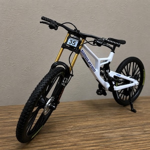 May include: A miniature white mountain bike model featuring gold-colored front suspension forks, black wheels with knobby tires, and a black handlebar. The frame displays the text SIMULATION and 14000, with a number 35 plate attached to the front.