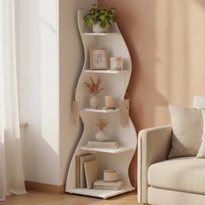 Op de afbeelding: Een witte, golvende hoekboekenplank met meerdere planken. De planken tonen een potplant, ingelijste kunst, kaarsen, vazen met gedroogde bloemen, boeken en macramé-versieringen. De kamer heeft een neutrale kleurenpalet.