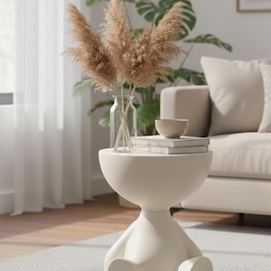 Puede incluir: Una mesa auxiliar de color crema con un diseño único, con una parte superior redondeada y una base con forma de figura sentada. La mesa contiene un jarrón de vidrio transparente con hierba de la pampa seca de color marrón, un cuenco pequeño y una pila de libros. La mesa está sobre una alfombra gris.