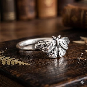 May include: A silver ring featuring a detailed moth design. The ring's band is smooth, leading to the intricately carved moth, showcasing patterns on its wings and body. The ring is set against a dark wooden surface.