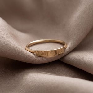 Puede incluir: Un anillo dorado con una placa rectangular grabada con las iniciales "MJ", seguido de un símbolo de corazón y la inicial "R". El anillo se encuentra sobre un tejido suave de tono neutro.
