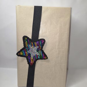 Op de afbeelding: Een cadeau verpakt in bruin papier, vastgezet met een zwart lint en een pailletten ster versiering. De ster heeft een regenboog aan kleuren en een zilveren centrum, wat een feestelijke touch toevoegt.