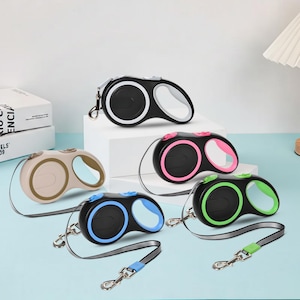 Op de afbeelding: Een verzameling intrekbare hondenriemen in verschillende kleuren, waaronder zwart, wit, roze, groen en blauw. Elke riem heeft een bijpassende handgreep en een reflecterende riem voor extra veiligheid tijdens wandelingen. De riemen zijn ontworpen voor kleine tot middelgrote honden.