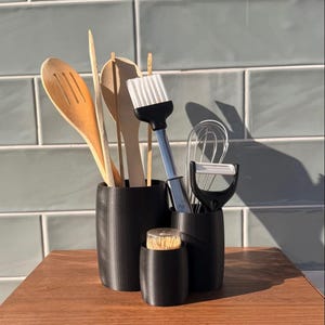 Peut inclure: Divers ustensiles de cuisine, dont des cuillères en bois, un fouet et un éplucheur, sont présentés dans trois supports cylindriques noirs. Les ustensiles sont disposés sur une surface en bois devant des carreaux bleu clair.