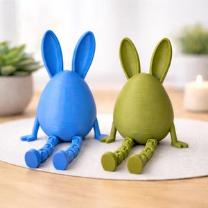 Könnte beinhalten: Zwei 3D-gedruckte Hasenfiguren. Eine ist blau, die andere grün. Jede hat einen eiförmigen Körper, lange Ohren und bewegliche Beine, was ihnen eine entspannte Pose verleiht. Diese dekorativen Artikel sind ideal für Ostern oder Frühlingsdekoration. Die Figuren sind ca. 10 cm hoch.
