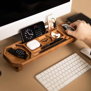 Puede incluir: Un organizador de escritorio de madera con múltiples compartimentos para accesorios. Contiene un teléfono inteligente con calendario, un estuche de auriculares blanco, un par de auriculares blancos, dos bolígrafos negros, un reloj y una foto.