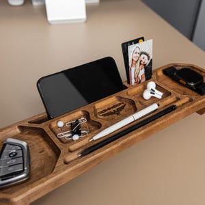 Op de afbeelding: Een houten bureau-organizer met meerdere vakken voor een smartphone, autosleutel, zwart-witfoto, witte draadloze oordopjes, een witte pen, een zwart potlood, paperclips en een smartwatch. Het hout is voorzien van de tekst JOHNSON.