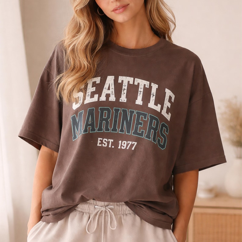 May include: A brown t-shirt with the text "SEATTLE MARINERS EST. 1977" in white and teal lettering. The shirt has short sleeves and a relaxed fit. The shirt is worn by a person.