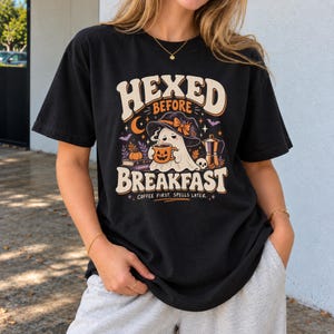 Op de afbeelding: Een zwart t-shirt met een afbeelding van een geest met een heksenhoed die een pompoenmok vasthoudt, samen met een koffiezetapparaat, boeken en vleermuizen. De tekst luidt "HEXED BEFORE BREAKFAST" en "COFFEE FIRST. SPELLS LATER." in retro letters.