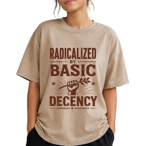 Op de afbeelding: Een beige t-shirt met korte mouwen en donkerbruine tekst met de tekst RADICALIZED BY BASIC DECENCY, met een afbeelding van een opgeheven vuist die een takje vasthoudt en de woorden KINDNESS RESISTANCE onderaan, gecentreerd op de borst.