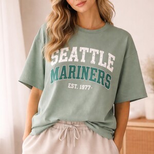 May include: Sage green t-shirt with the text "SEATTLE MARINERS" in white and teal lettering. The text "EST. 1977-" is below the team name. The shirt has short sleeves and a relaxed fit.