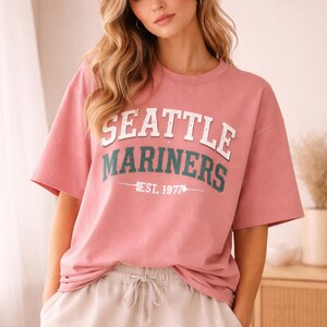 May include: A dusty rose-colored t-shirt with the words "SEATTLE MARINERS" in white block letters and teal lettering, with "EST. 1977" below. The shirt has short sleeves and a crew neck.