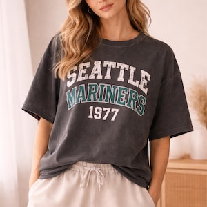 May include: Dark gray t-shirt with the text "SEATTLE MARINERS 1977" in white and teal lettering. The shirt has short sleeves and a relaxed fit. The model is wearing light-colored pants.