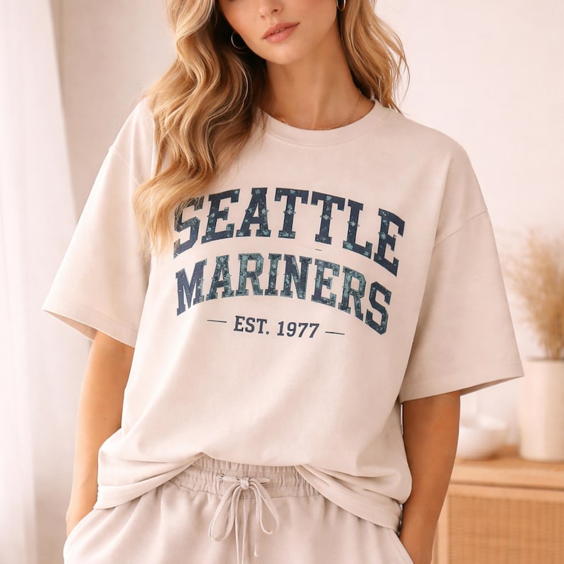 May include: Cream-colored t-shirt with the text "SEATTLE MARINERS" in navy blue, with a patterned design, and the year "EST. 1977". The shirt has short sleeves and a crew neck. The person is wearing matching cream-colored sweatpants.