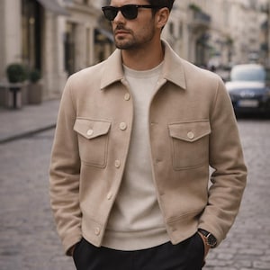 May include: A beige jacket with two front pockets and button closures. The jacket is worn over a cream-colored sweater. The person is wearing black sunglasses and black pants. A watch is visible on the wrist.