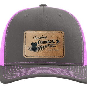 Op de afbeelding: Grijze truckerpet met een roze mesh achterkant en klep. Een rechthoekige patch toont de woorden "Traveling Courage Project" met een hart- en pijldesign. De pet is een klassieke stijl, geschikt voor vrijetijdskleding.