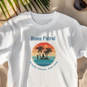 May include: White t-shirt with a circular graphic featuring a beach scene with palm trees, surfers, and a sunset. Text on the shirt reads "Dawn Patrol" and "Empty waves, Full soul."