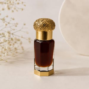 May include: A small, dark brown perfume bottle with a gold filigree cap and gold accents. The bottle has a geometric base and a cylindrical body. The background is a soft, neutral colour, with a sprig of white flowers.