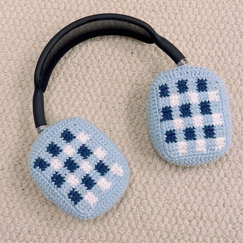 May include: Over-ear headphones with light blue crochet ear covers featuring a checkered pattern in white and navy blue. The headband is black with a padded top. The earcups are oval-shaped.