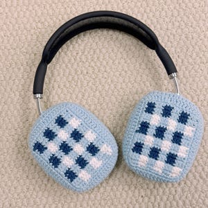 May include: Over-ear headphones with light blue crochet ear covers featuring a checkered pattern in white and dark blue. The headband is black with silver-colored metal arms.