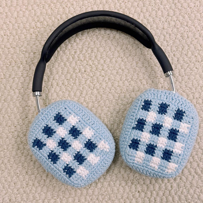 May include: Over-ear headphones with light blue crochet ear covers featuring a checkered pattern in white and dark blue. The headband is black with silver-colored metal arms.
