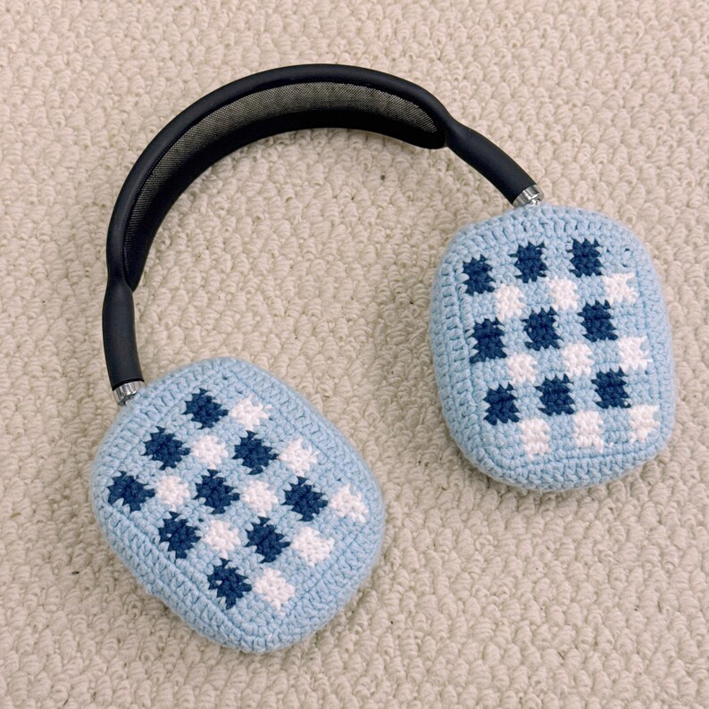 May include: Light blue headphones with a black headband. The earcups are covered in a crocheted pattern of white and navy blue squares. The headphones are resting on a light-colored, textured surface.