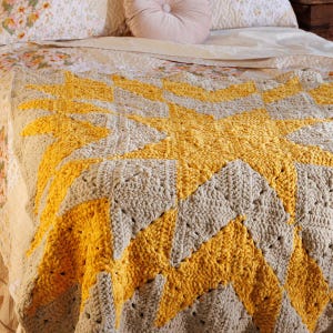Könnte beinhalten: Eine grob gehäkelte Decke mit einem geometrischen Sternmuster aus abwechselnd senfgelbem und hellgrauem Garn. Das Textil weist eine dicke, strukturierte Maschenkonstruktion mit einem sich wiederholenden kantigen Design auf der gesamten Fläche auf.
