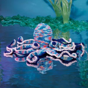 Könnte beinhalten: Ein gehäkeltes Oktopus-Spielzeug mit einem mehrfarbigen Garnmuster in Blau-, Rosa- und Weißtönen. Der Oktopus hat einen abgerundeten Kopf und acht gewellte Tentakel, die jeweils eine einfarbige dunkelblaue Unterseite und einen weißen Häkelrand haben.