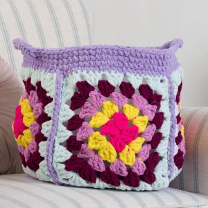 Könnte beinhalten: Ein quadratischer Häkelkorb aus mehreren Granny Squares mit einer lebendigen Farbpalette aus lavendelfarbenem, hellblauem, dunkelburgunderrotem, leuchtend gelbem und pinkfarbenem Garn. Der obere Rand ist mit einer dicken lavendelfarbenen Häkelkante versehen.