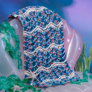 May include: A chunky crochet blanket featuring a variegated yarn pattern in shades of blue, teal, and coral, accented with horizontal white wavy lines. The blanket is decorated with two small, light-colored starfish placed on the textured surface.