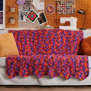 Könnte beinhalten: Eine grob gehäkelte Decke mit einem lebendigen, mehrfarbigen Garnmix aus leuchtendem Pink, hellem Blau, Orange und Gelb. Die Decke weist ein strukturiertes Maschenmuster auf und liegt über einem hellen Sofa mit zwei einfarbigen Kissen.