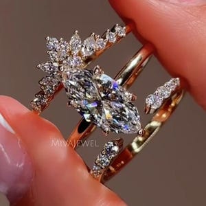 Peut inclure: Ensemble de bagues en or ornées de diamants étincelants. La bague principale présente un diamant taille marquise, complétée par un anneau fin et une bague en forme de chevron. Les bagues sont tenues par une main.