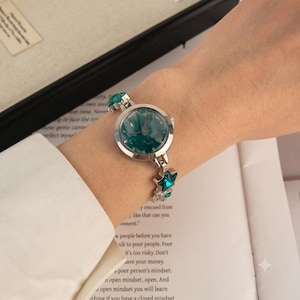 Peut inclure: Une montre analogique argentée dotée d'un cadran rond au motif marbré vert foncé avec des aiguilles argentées. Le bracelet se compose de maillons argentés ornés de petits cristaux vert foncé en forme d'étoile et de pierres transparentes.
