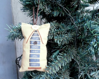 May include: A handmade fabric ornament shaped like a house with a blue and white striped design. The ornament is hanging on a green artificial Christmas tree.